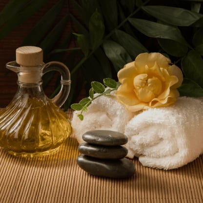 Massageöl, weiße Handtücher, eine gelbe Blüte und Massagesteine auf einer Bambusmatte